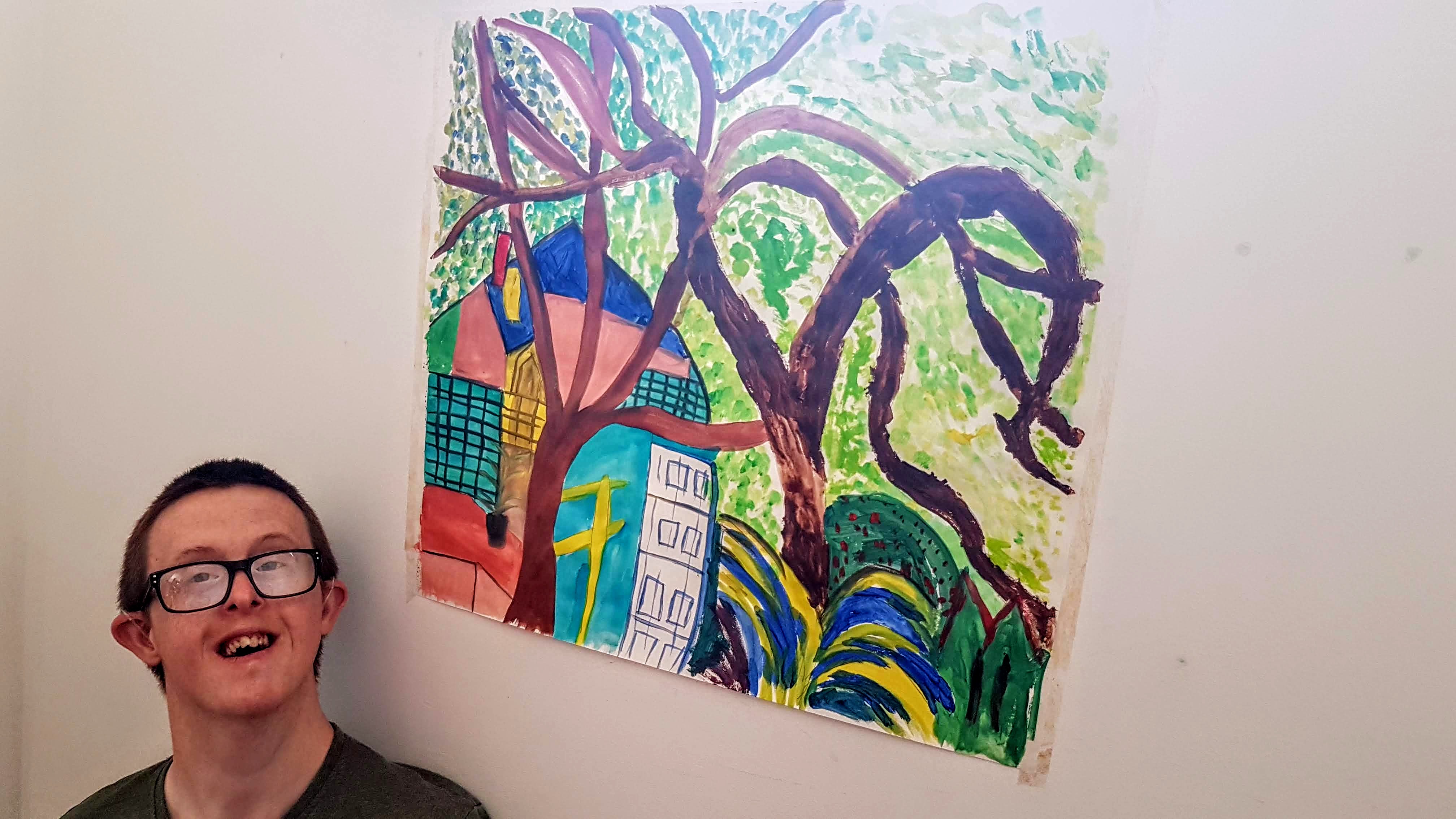 Young adult headshot looking to camera with his artwork in background, a house, tree and leaves painted in vibrant colours